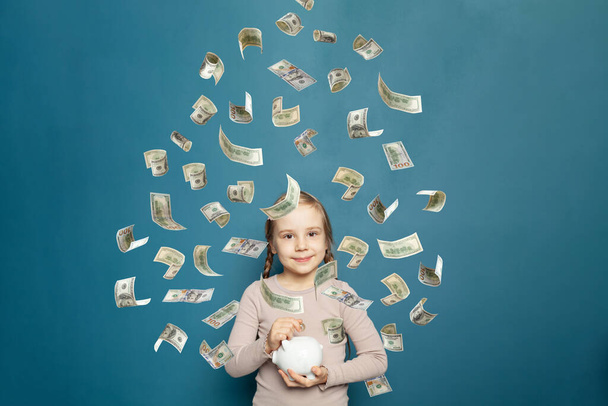 Beautiful young child girl with flying money on blue background - Photo, Image