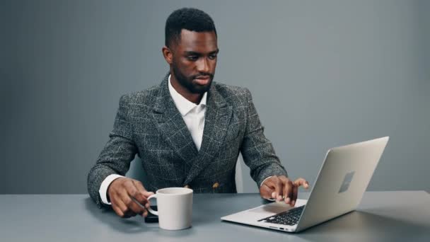 Gelukkige Afro-Amerikaanse man werkt op een laptop in het kantoor aan zijn bureau drinken verkwikkende koffie en glimlachen. Hoge kwaliteit 4k beeldmateriaal - Video