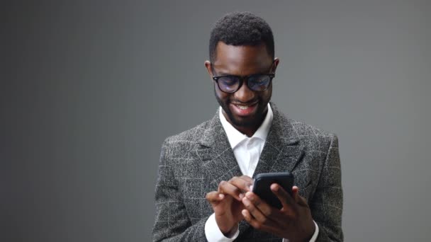 Afrikai-amerikai üzletember kezében egy telefont néz rá, és boldog, hogy nyerni és nyerni az irodában szürke alapon. Kiváló minőségű 4k felvételek - Felvétel, videó