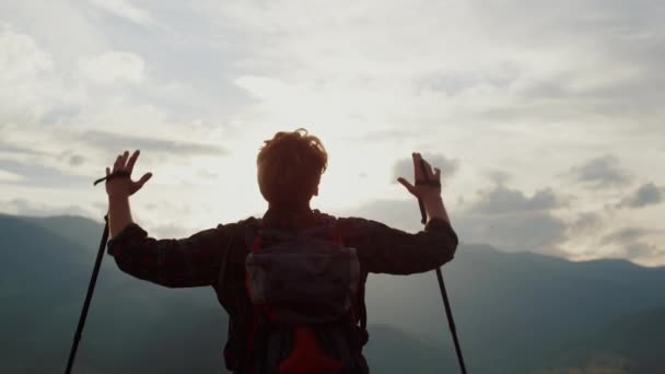 Joyful hiker raise hands to mountains sky. Unknown tourist silhouette celebrate freedom on hill peak close up. Back view of man hold trekking poles on adventure. Traveler enjoy nature. Happy concept. - Video