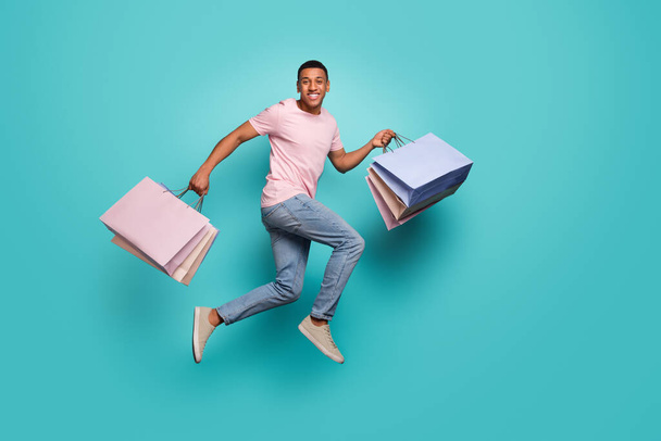 Full length photo of charming impressed guy wear pink t-shirt jumping high holding shoppers empty space isolated teal color background. - Zdjęcie, obraz
