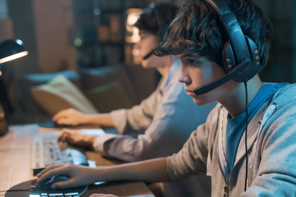 Teenager friends playing online video games together, they are wearing headsets and staring at the computer monitor - Foto, Imagem