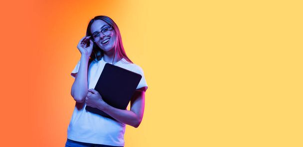 Enjoying student life. Portrait of young smiling girl in white t-shirt with laptop isolated on orange color background in neon light. Concept of beauty, art, fashion, education - 写真・画像
