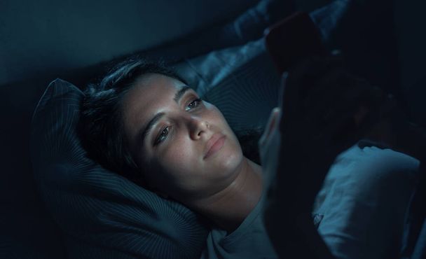 Young woman lying in her bed at night and connecting with her smartphone, she can't sleep - Foto, imagen