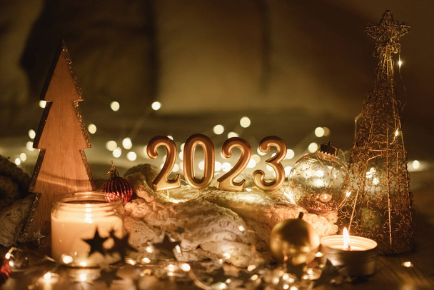 Happy New Year 2023. Number 2023 made by candles on a festive sparkling bokeh background in the dark. New Year background with sweaters, garlands and New Year decorations. - Photo, Image