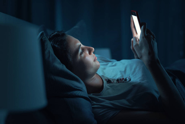 Young woman lying in her bed at night and connecting with her smartphone, she can't sleep - Fotó, kép