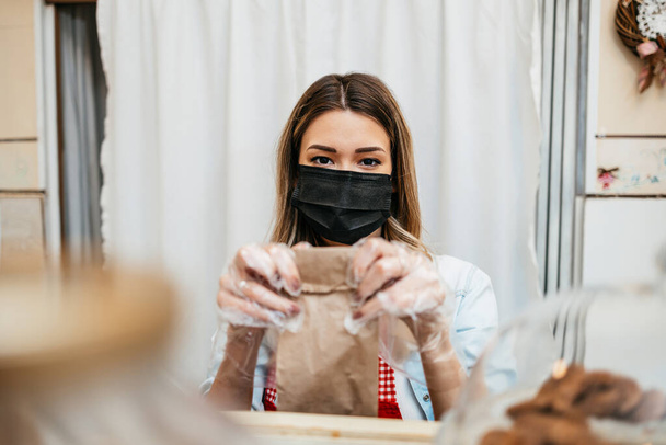 Beautiful young female seller with protective face mask working in a confectionery shop or bakery. She is selling and packing delicious sweets. Coronavirus and small business concept. - Фото, изображение