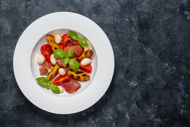 Grilled peach salad with mozzarella, prosciutto ham, red tomato, green basil and soy sauce. Fresh gourmet salad in white plate on black background, close up, top view, copy space - Photo, Image