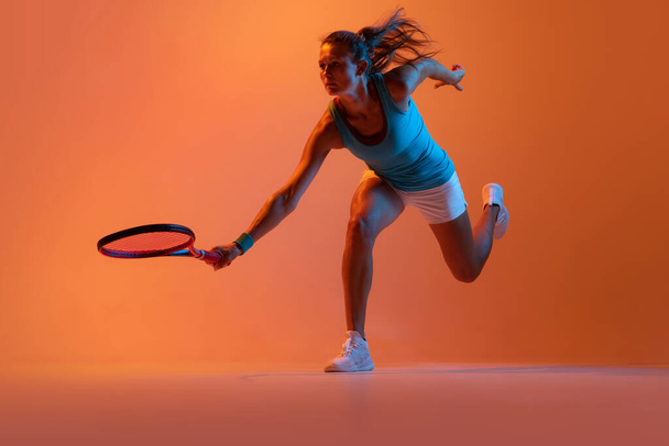 Dynamic portrait of woman, professional tennis player training isolated over orange background in neon light. Concept of sport, health, strength, action, motion, hobby, lifestyle. Copy space for ad - Fotoğraf, Görsel