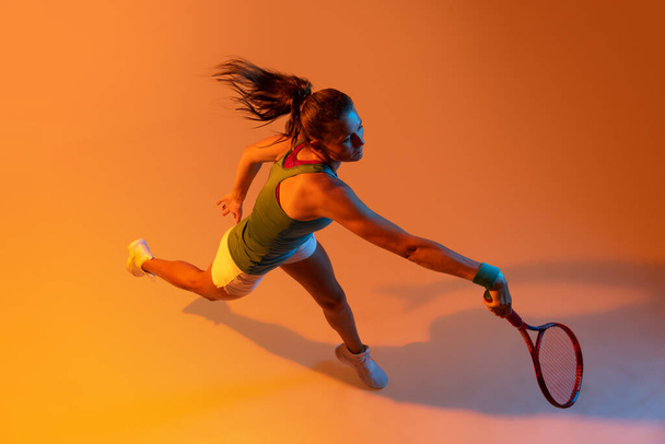 Top view portrait of woman, professional tennis player training isolated over orange studio background in neon light. Concept of sport, health, strength, motion, hobby, lifestyle. Copy space for ad - Foto, Imagem