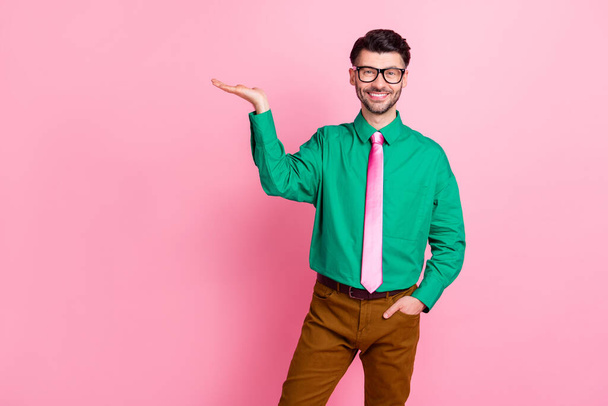 Portrait of young stylish guy wear green shirt pink tie holding hand new product advertisement isolated on pink color background. - Photo, Image