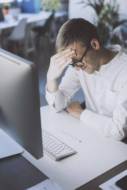 Man sitting at desk and working with his computer, he is tired and he has a bad headache - Фото, изображение