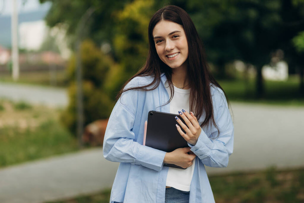 portrait of a beautiful student girl. Student in green grass, with backpack, smiling, holding tablet in hands - Foto, afbeelding