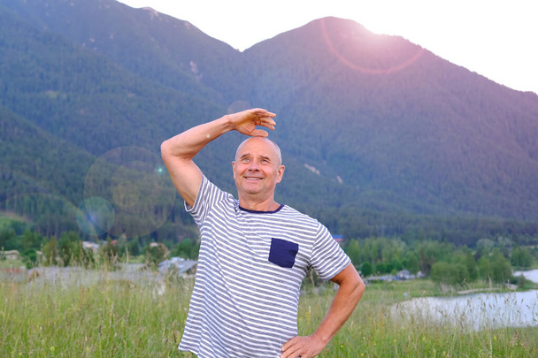 mature man, senior 60 years old standing in mountains above city, smiles, Alps are behind him, green grass in meadow, concept of enjoy life in old age, travel, active lifestyle, human happiness - Photo, image