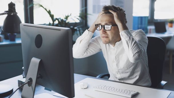Panicked businessman sitting at desk and staring at the computer screen, system failure and computer problems concept - Photo, image