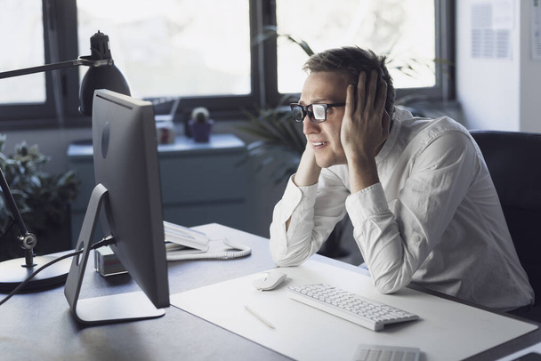 Panicked businessman sitting at desk and staring at the computer screen, system failure and computer problems concept - 写真・画像