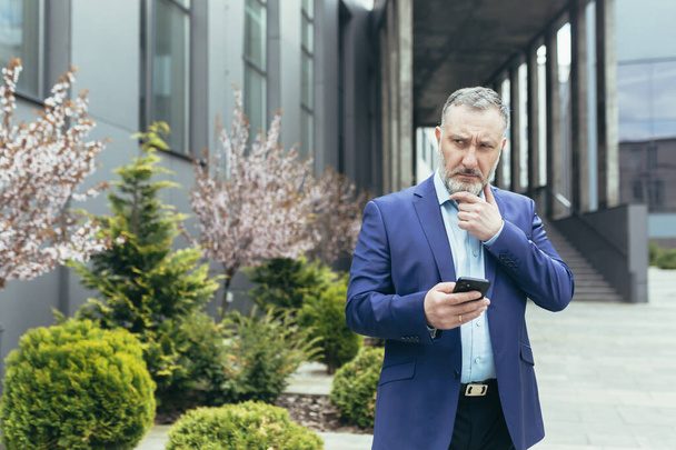 pensive senior and experienced man outside office reading news on phone, businessman outside office using smartphone while walking near building - Foto, immagini
