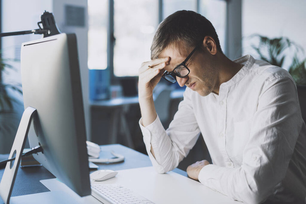 Man sitting at desk and working with his computer, he is tired and he has a bad headache - Photo, image