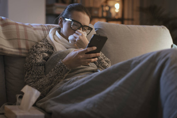 Sick young woman resting on the couch at home and connecting online using her smartphone, telemedicine concept - Foto, imagen