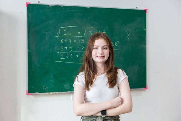 Girl is standing in a classroom with a green chalkboard covered with formulas and figures. Kid write on chalk board. - Foto, Imagem