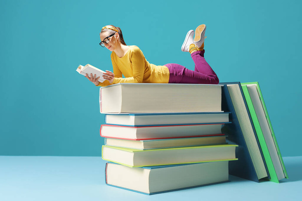 Tiny smart woman lying on a pile of books and reading, education and hobby concept - Foto, afbeelding