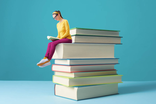 Tiny smart woman sitting on a pile of books and reading, education and learning concept - Фото, изображение