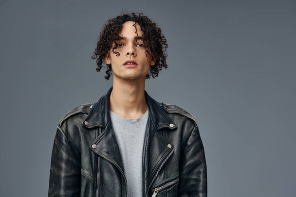Portrait of serious stylish tanned curly man leather jacket looks at camera posing isolated on gray studio background. Cool fashion offer. Huge Seasonal Sale New Collection concept. Copy space for ad - Photo, Image