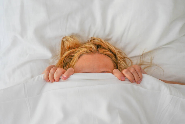 Woman lying in bed and hiding her face behind sheet. Woman with stressed face expression hiding behind sheet in bed. Concept of avoiding responsibility - Photo, image