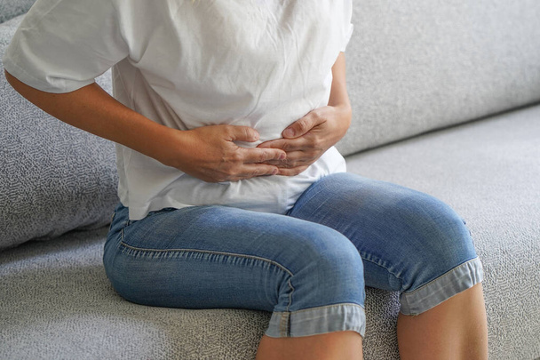 Woman siting on sofa and holding her hands on her stomach. Woman having painful stomachachem, menstruation. Pain in the abdomen of a woman. Body and health care concept - Photo, image
