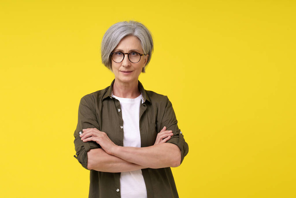 Mature woman in her 50s, serious businesswoman with folded hands looking straight in camera wearing green shirt with white t-shirt isolated on yellow background. Mature people beauty concept. - Foto, immagini