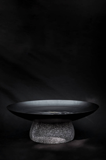 bowl with metal water and natural stone base, works as a water mirror, decoration, design. Black background with natural filtered light, the stone is terrazzo, quarry or recinto. Works as a pot or - Photo, image