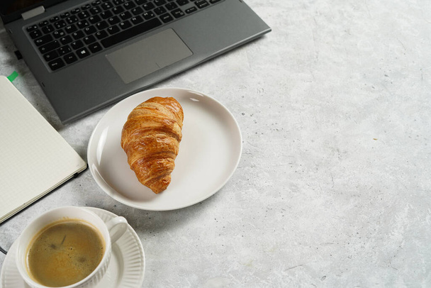 A croissant and a cup of coffee on white plates on grey working desk in the office with laptop and notebook, copy space for text - Photo, image