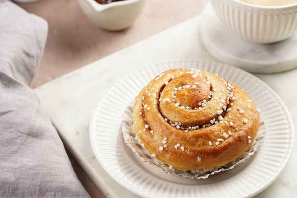 A round cinnamon pastry on a white plate on marble board on light pink background, cup of coffee, jam, milk - Foto, Imagen