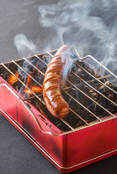 A fresh pork sausage grilling over an improvised barbecue on a metal box. - Zdjęcie, obraz