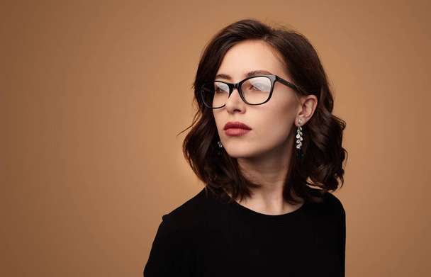 Confident young female model with dark wavy hair and makeup in elegant clothes and eyeglasses looking away against beige background - Foto, Bild