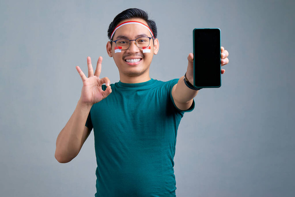 Smiling young Asian man in casual t-shirt showing blank screen mobile phone and gesturing okay sign isolated on grey background. indonesian independence day celebration concept - Photo, image