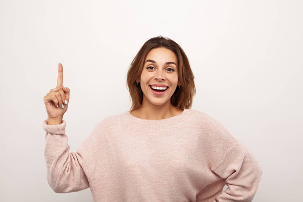 Optimistic young woman with long brown hair in casual clothes smiling widely and pointing up while standing against white background with hand on waist - Foto, immagini
