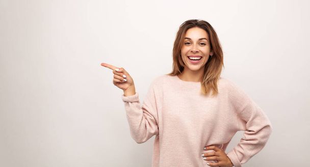 Confident young woman with brown hair in casual outfit pointing aside and smiling at camera while standing in studio with hand on waist - Foto, Bild