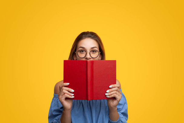 Smart young female with long brown hair in blouse and eyeglasses covering mouth with red book and looking at camera against yellow background - Valokuva, kuva