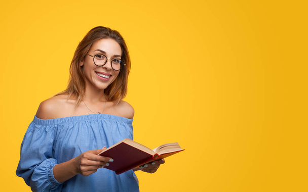 Positive young smart woman with long brown hair in stylish blouse and eyeglasses, smiling and looking at screen while reading interesting book against bright yellow background - Foto, afbeelding
