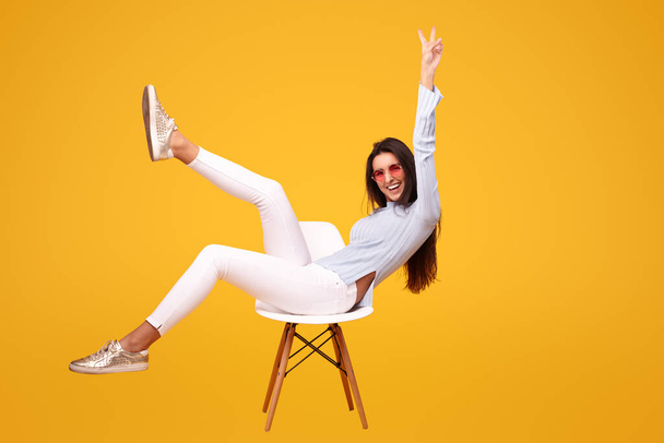 Side view of excited young ethnic female millennial in casual wear and sunglasses, sitting on chair with raised legs while smiling and showing V sign against yellow background - Fotó, kép