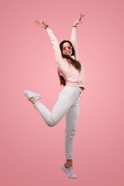 Side view full body of excited young female model with long dark hair in stylish clothes and sunglasses, smiling and raising arms while standing against pink background - Zdjęcie, obraz