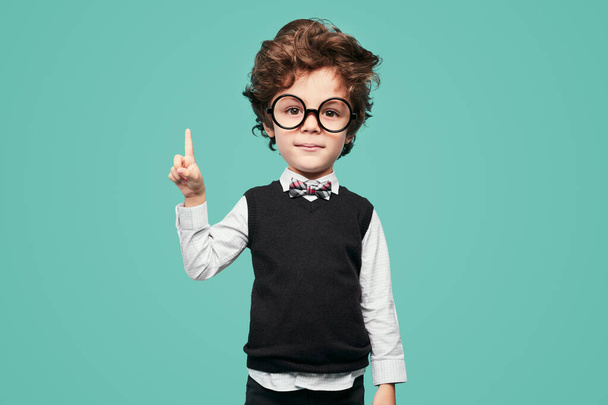 Cute clever little schoolboy with big had and brown hair in classy clothes with bow tie and round eyeglasses, pointing up and looking at camera against turquoise background - Fotoğraf, Görsel