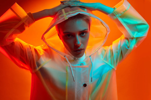 Unemotional young female model with short blond hair in trendy transparent hooded raincoat, touching head and looking at camera against orange background in studio with blue neon light - Fotó, kép