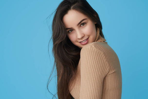 Side view of self assured young female model with long dark hair in beige turtleneck, smiling and looking at camera with confidence against blue background - Foto, Bild