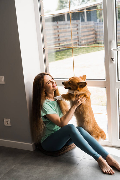 Happy girl hug Corgi dog at home. Lifestyle with domestic pet. Young woman sitting and play with Welsh Corgi Pembroke - Photo, Image