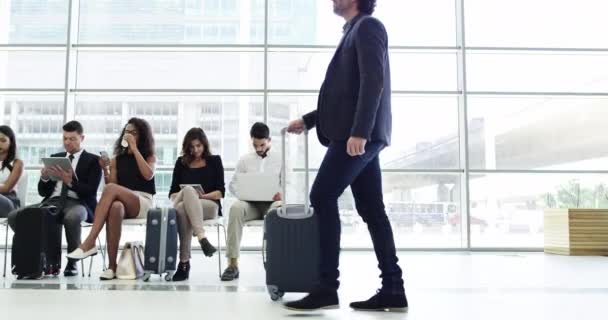 Молоді бізнесмени в аеропорту чекають свого польоту, сидять у вестибюлі перед поїздкою і працюють над своїми цифровими гаджетами. Чоловік тягне свій проїзний багаж до воріт-інтернатів
. - Кадри, відео