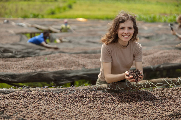 Smiling lady holding dried coffee beans in hands at farm in Africa region with workers on the background - Фото, зображення