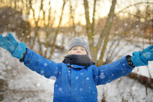 Portrait of funny little boy in blue winter clothes walks during a snowfall. Cute child wearing a warm clothing, hat, scarf. Outdoors winter activities for family with kids. The first snow - Φωτογραφία, εικόνα