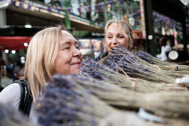 A senior woman is smiling a bush of lavender in a street market and her friend is looking to her having fun. Borough market, London. - Fotografie, Obrázek